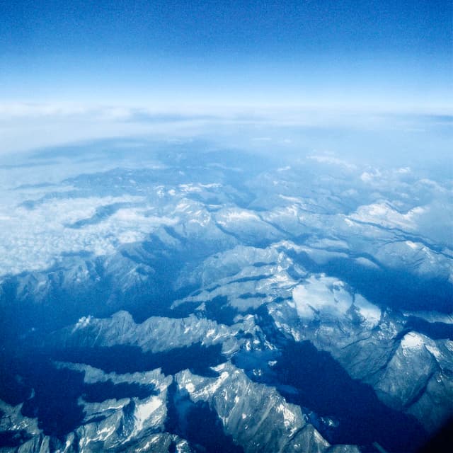 Aerial view of snow-capped mountain peaks under a clear blue sky, capturing a serene, expansive landscape with bright, natural daylight and shadows