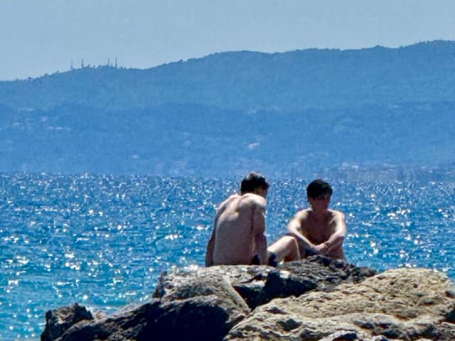 two shirtless men sit on dark coastal rocks under bright sunlight with blue ocean water and distant mountains in the background
