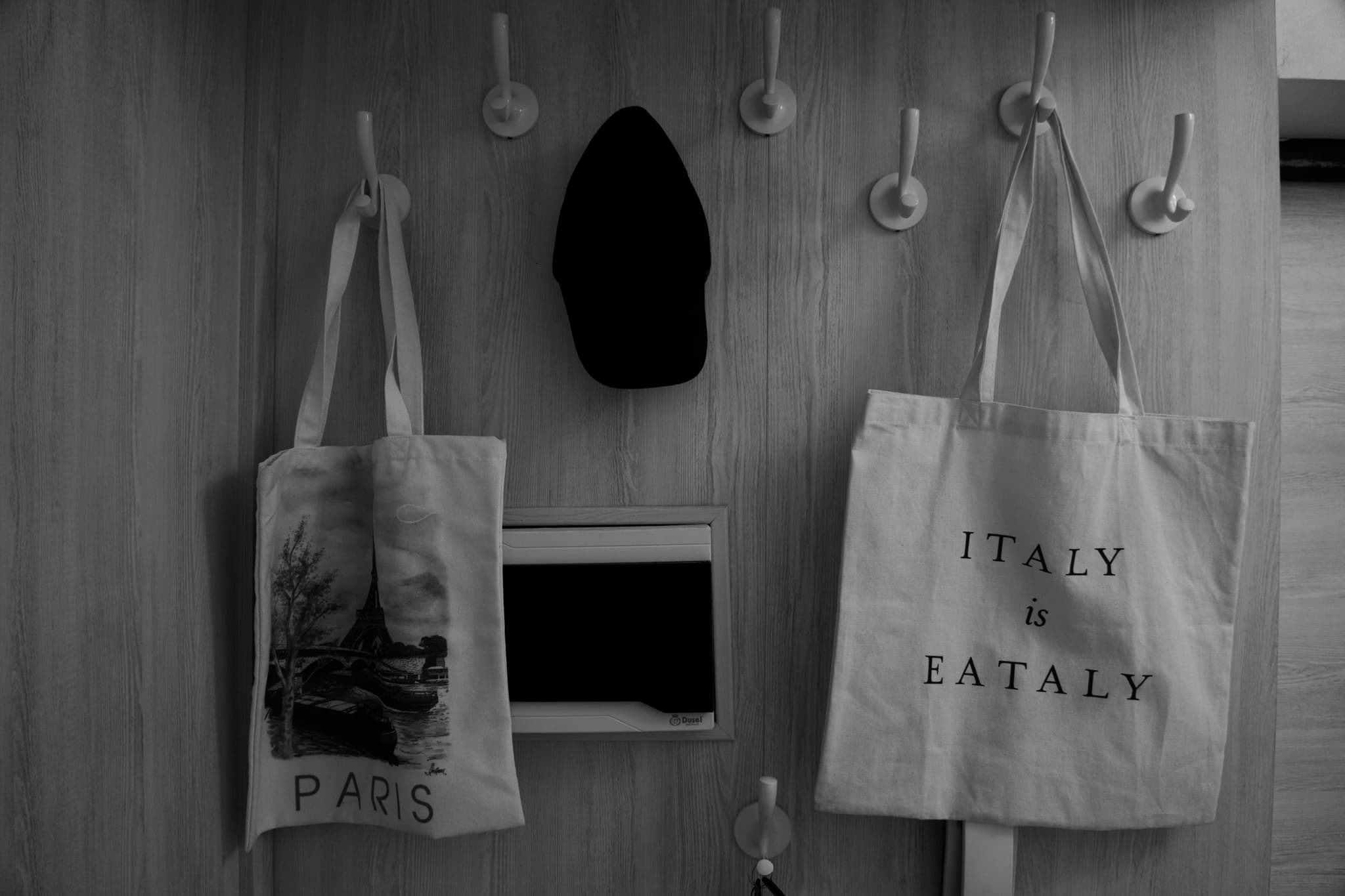 two canvas tote bags and a black cap hang from wall hooks in a dimly lit room, captured in high-contrast black and white