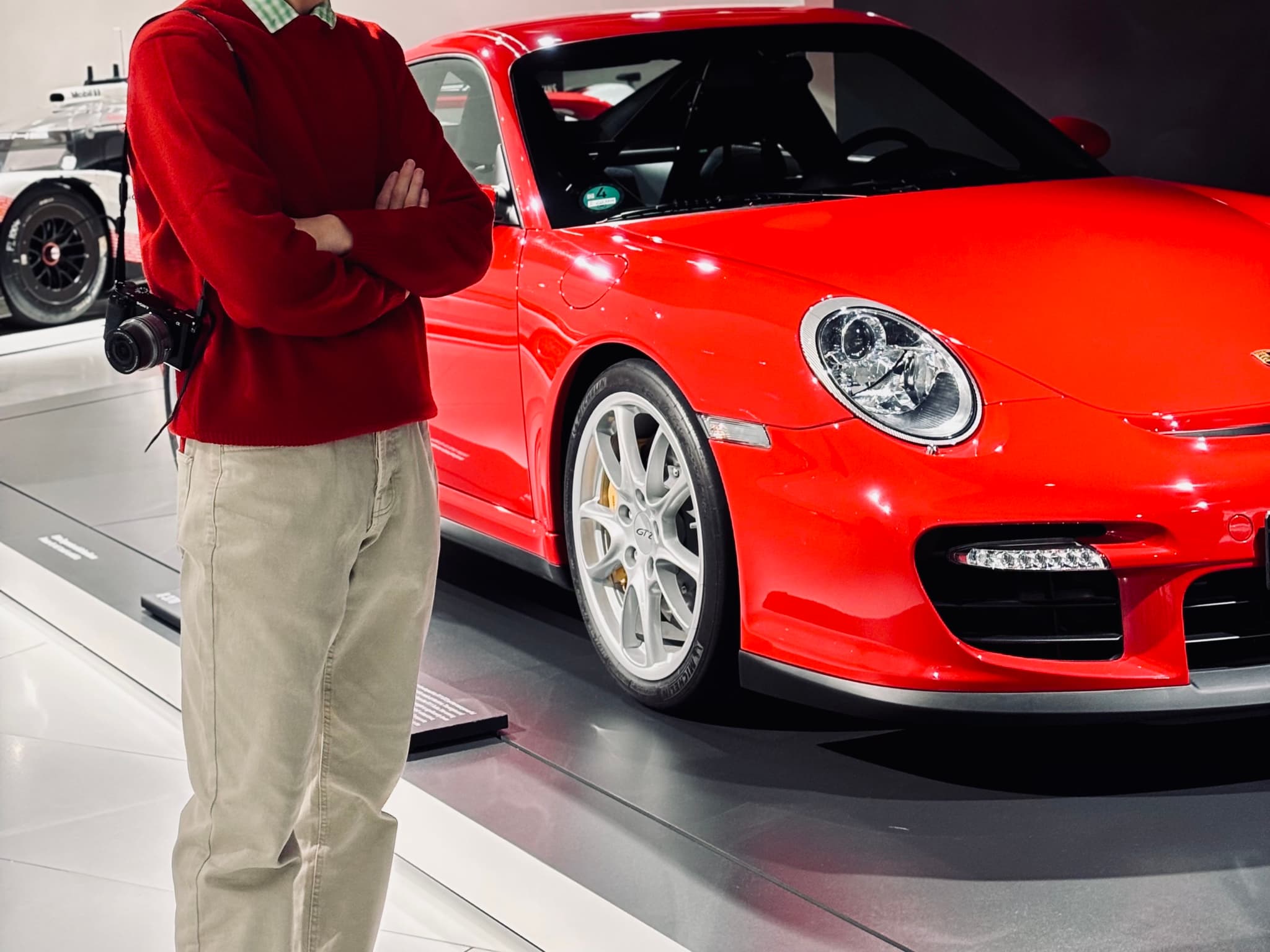 person in red sweater standing beside a bright red porsche sports car inside a white museum gallery under artificial indoor lighting