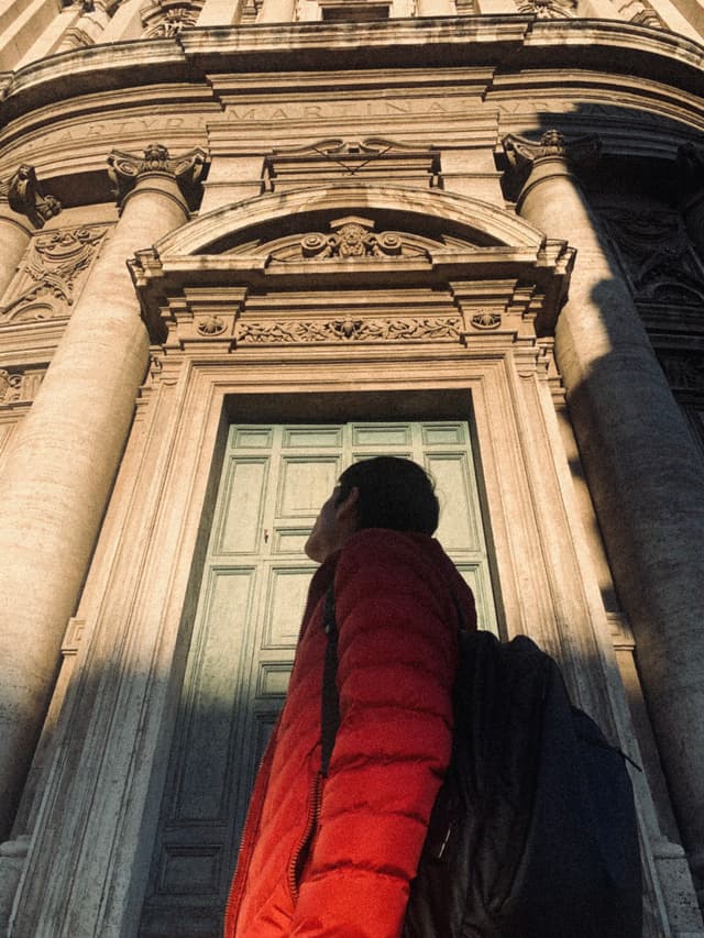 person in red jacket looks up at stone church entrance during late afternoon light in rome, capturing architectural scale and travel mood