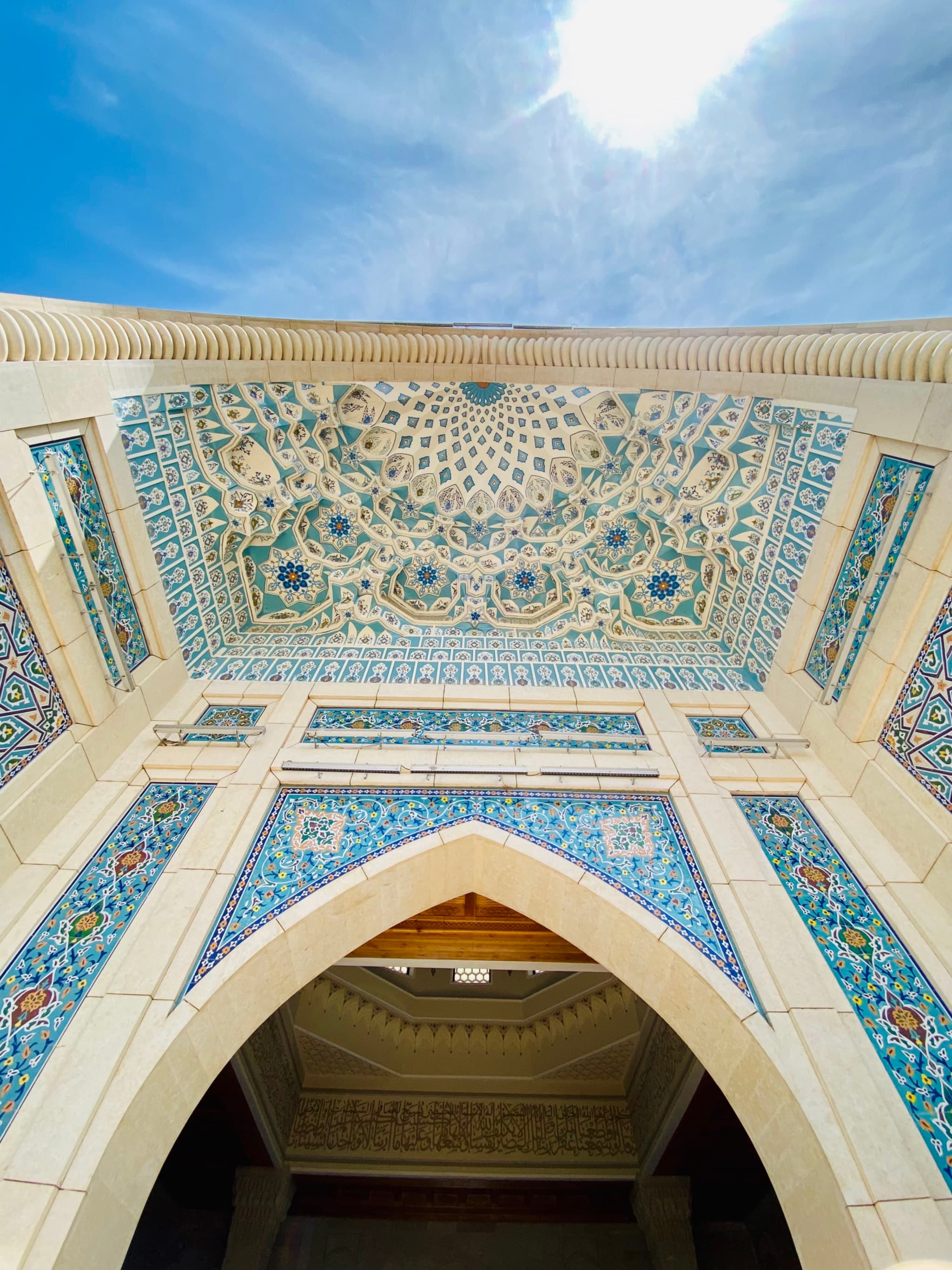 low angle view of a mosque entrance with blue tile mosaics and geometric patterns under a bright midday sun in clear sky