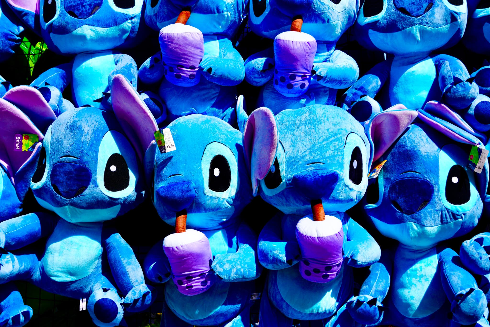 rows of blue stitch plush toys holding purple drinks under bright lighting, arranged in a repetitive display for a carnival game