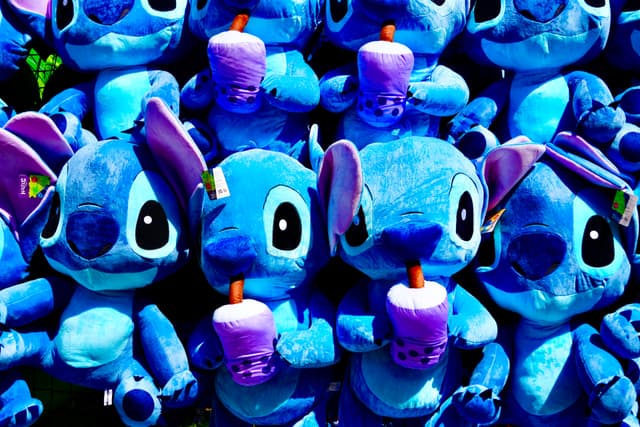 rows of blue stitch plush toys holding purple drinks under bright lighting, arranged in a repetitive display for a carnival game