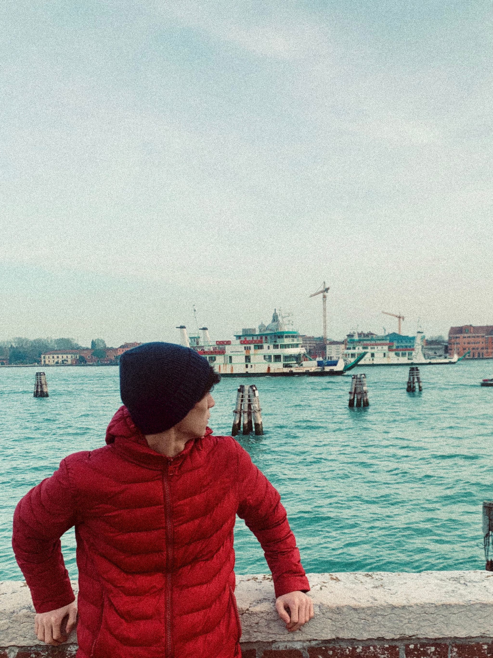 person in a red jacket and black beanie stands by the venice canal under flat daylight with a ferry in the background