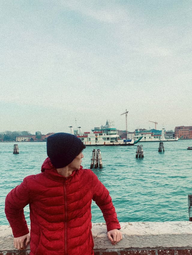 person in a red jacket and black beanie stands by the venice canal under flat daylight with a ferry in the background