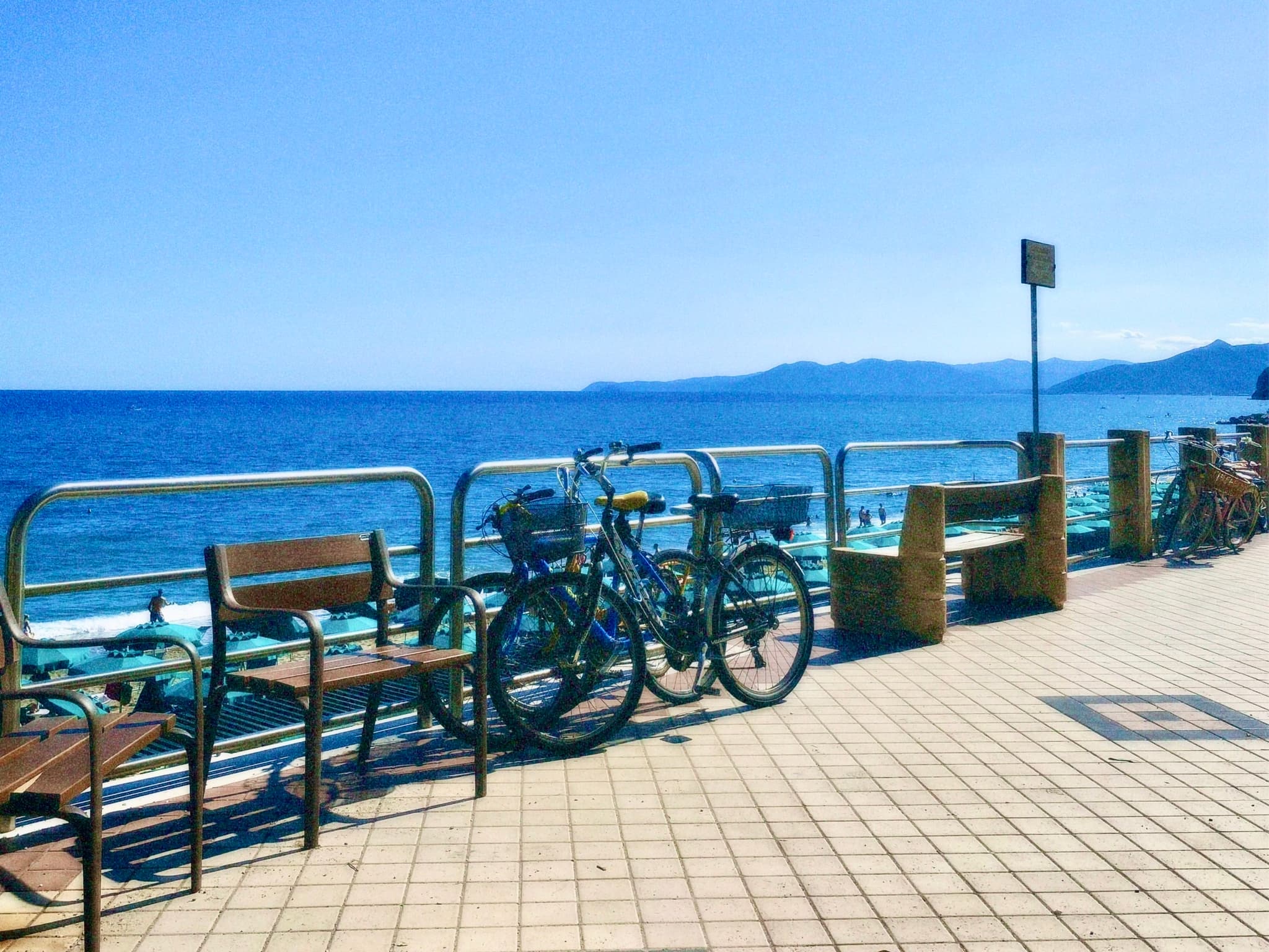 two bicycles parked on a tiled coastal promenade overlooking the blue sea under bright sunlight with wooden benches and metal railings