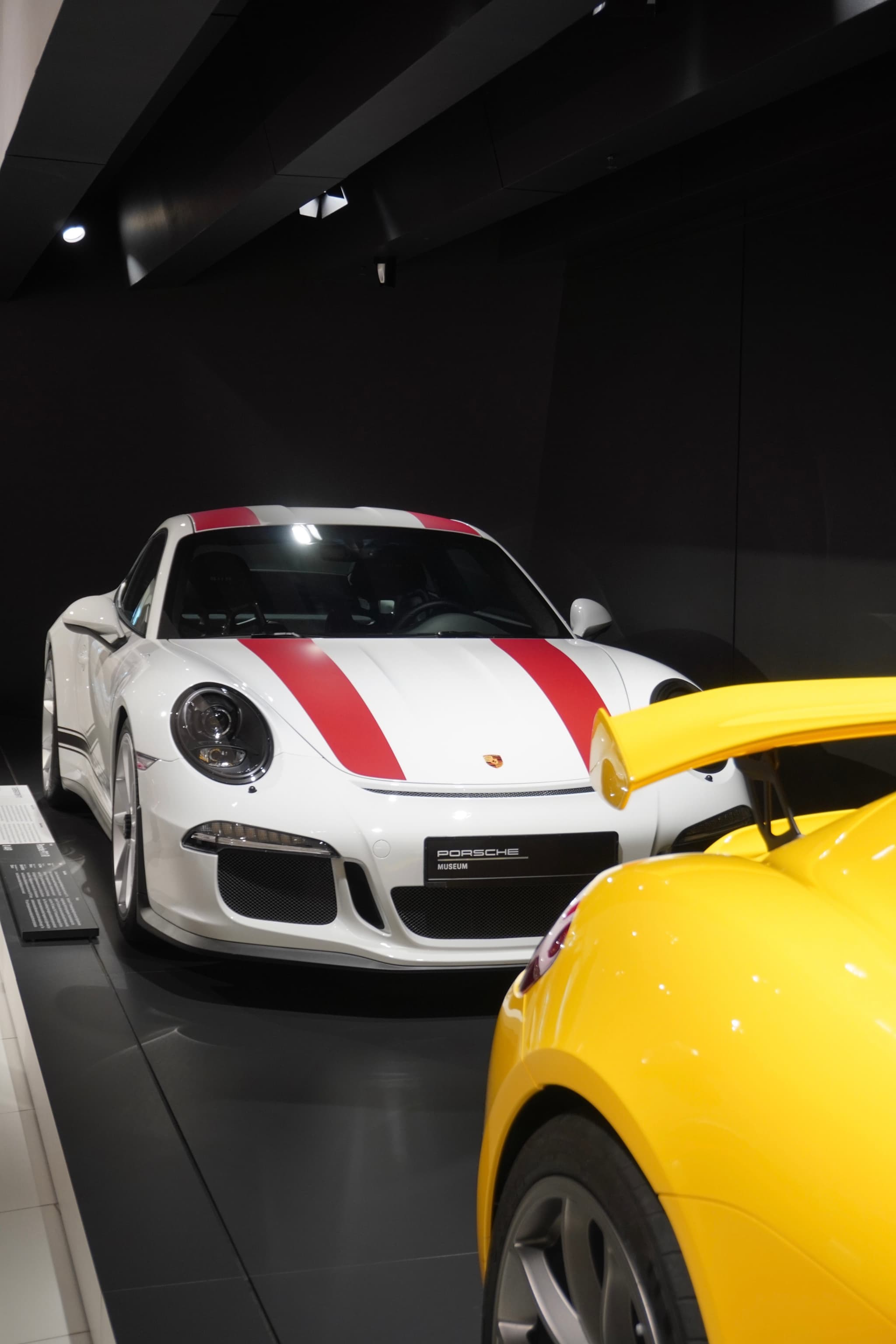 white porsche with red stripes parked behind a yellow sports car inside a dark museum gallery under bright overhead spotlights