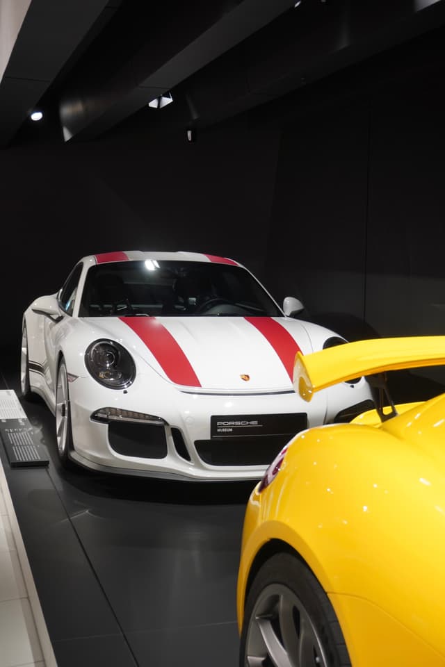 white porsche with red stripes parked behind a yellow sports car inside a dark museum gallery under bright overhead spotlights
