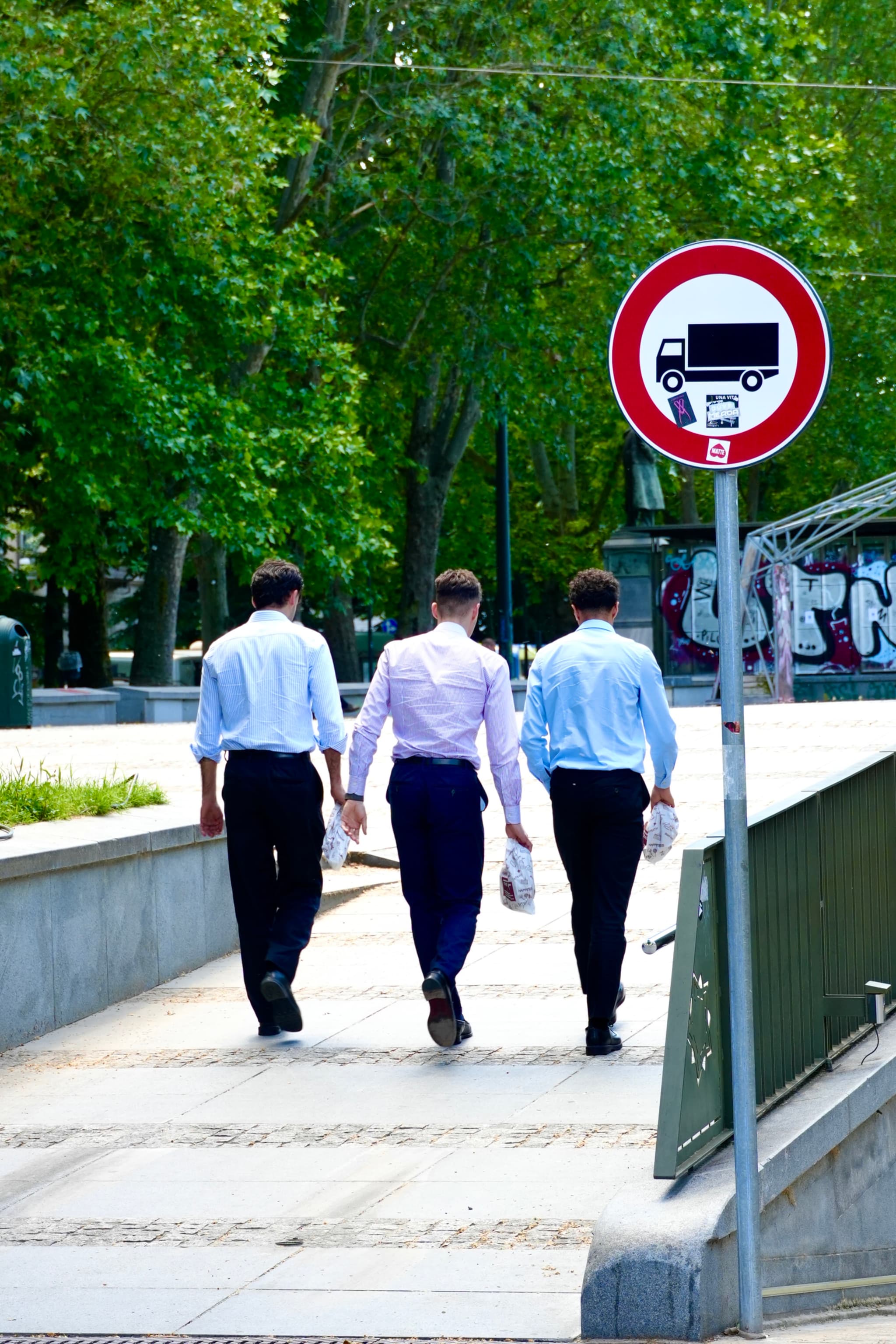 three men in dress shirts walk on a city sidewalk past a no trucks sign under bright daylight in a public park