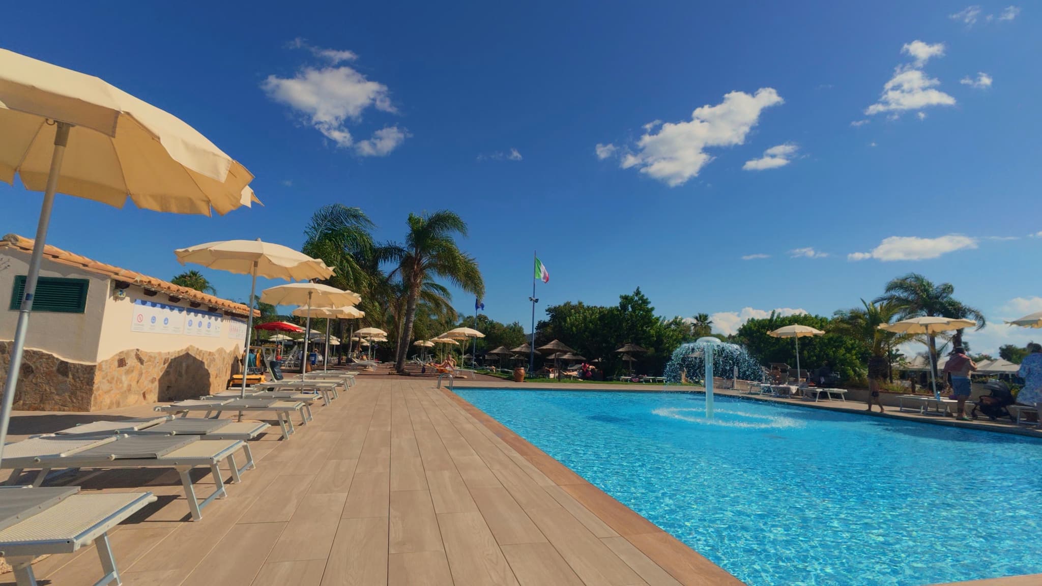 bright blue swimming pool with a central fountain, wooden deck, beige umbrellas, and palm trees under a clear sky on a sunny day