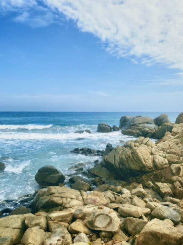 waves crash against large tan rocks on a sunny coastline under a bright blue sky with scattered white clouds at midday