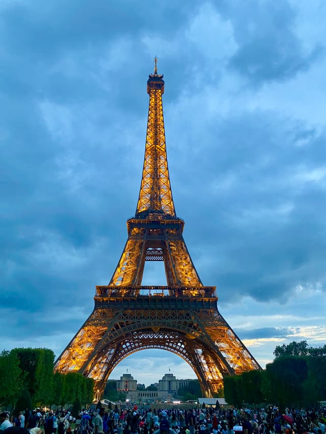 eiffel tower illuminated at dusk with golden lights against a cloudy blue sky, viewed from a crowded park in central paris, france