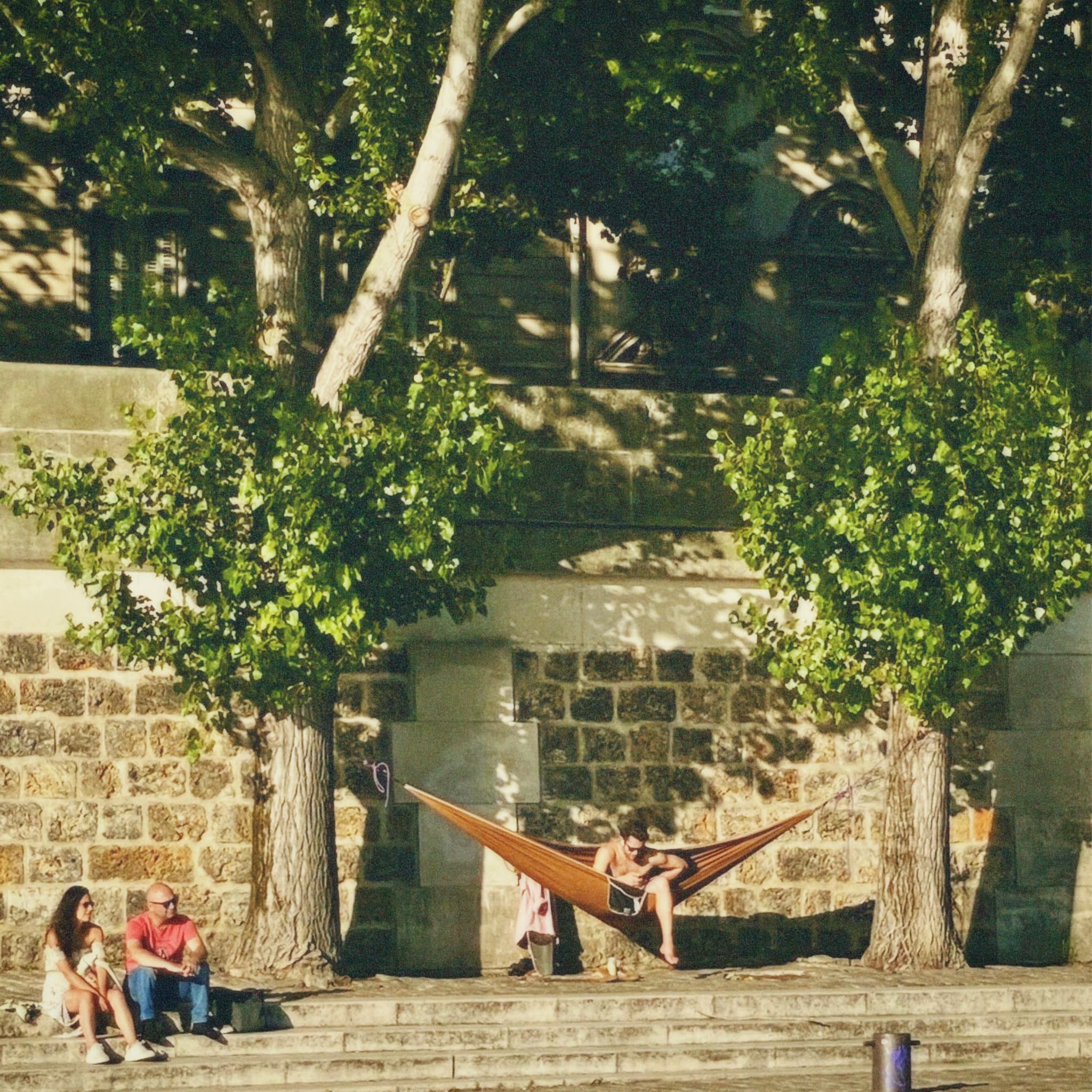 people relax in a hammock and on stone steps along the seine riverbank during a sunny afternoon with dappled tree shadows
