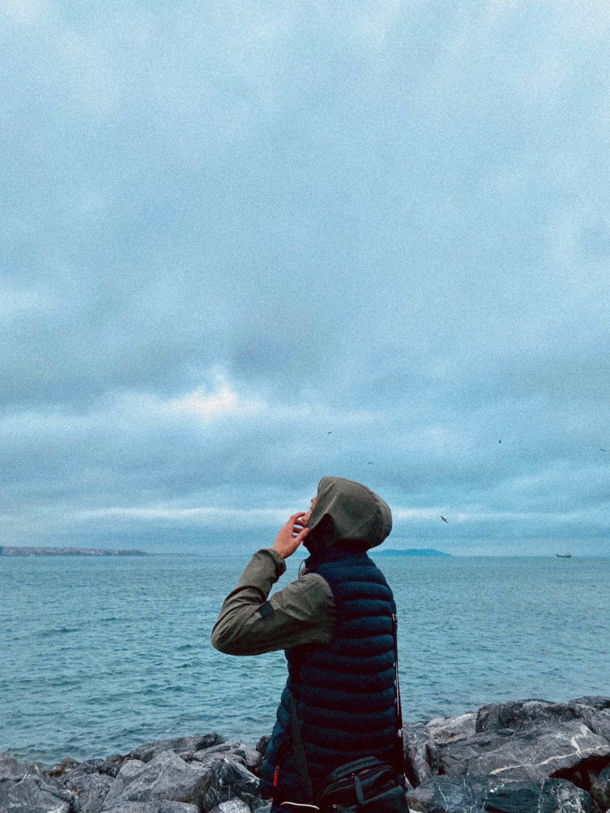 person in a hooded jacket stands on a rocky shore looking at a cloudy sky over the cold blue ocean water