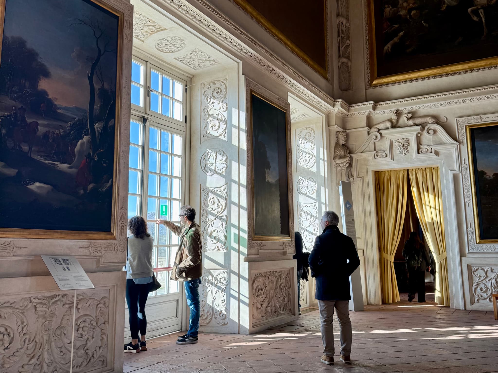 visitors view large oil paintings in a bright palace gallery with tall windows, ornate moldings, and soft afternoon light on wooden floors