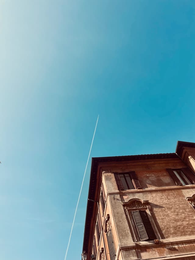 white vapor trail crosses a clear blue sky above the corner of a stone building in bright daylight, low angle view