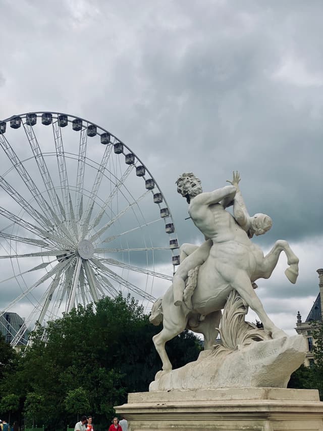 stone centaur statue stands before a large ferris wheel in a paris park under a cloudy sky during a flat afternoon