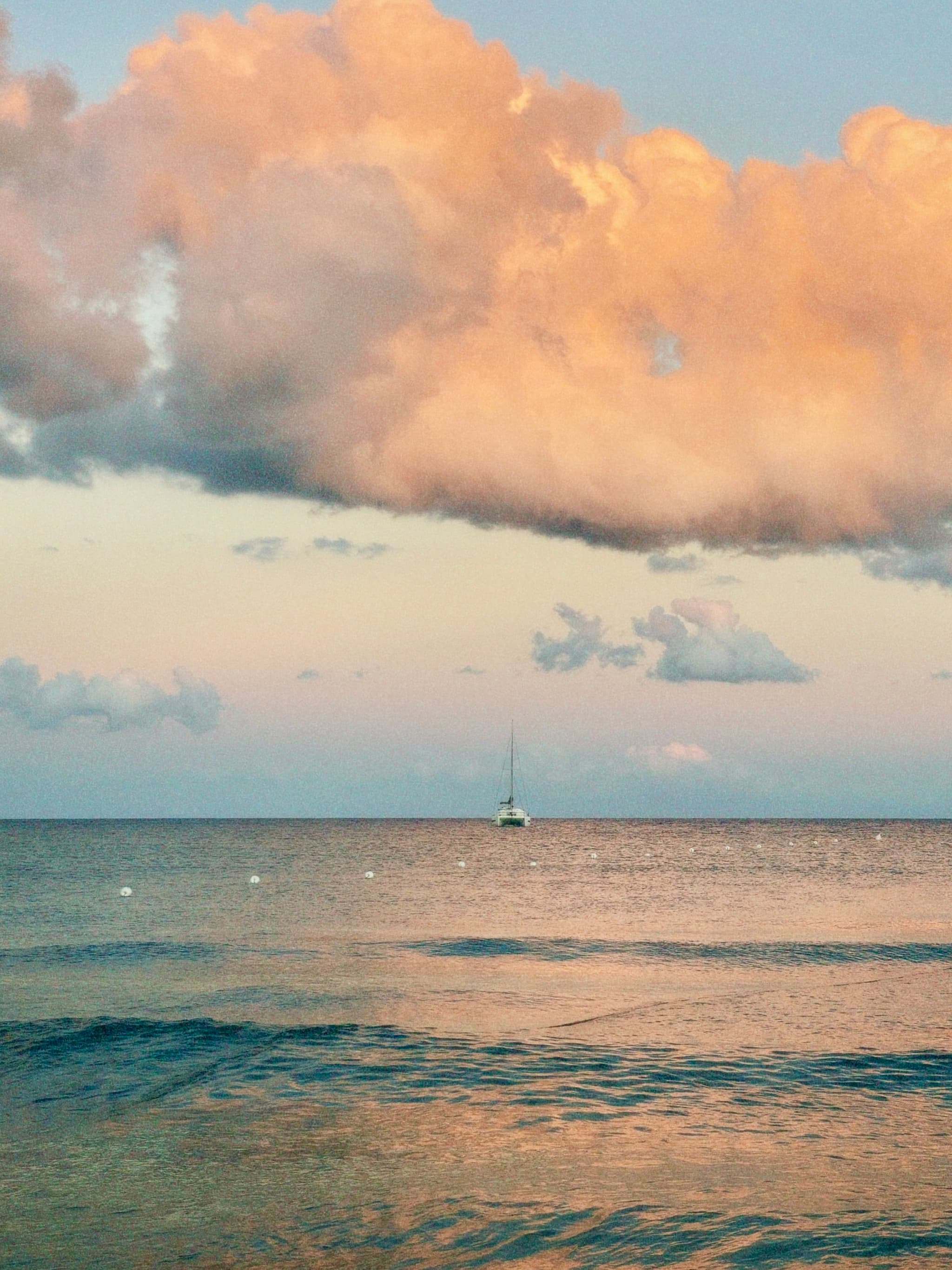a lone sailboat floats on calm water under pink clouds during sunset, captured with soft natural light in a quiet coastal setting