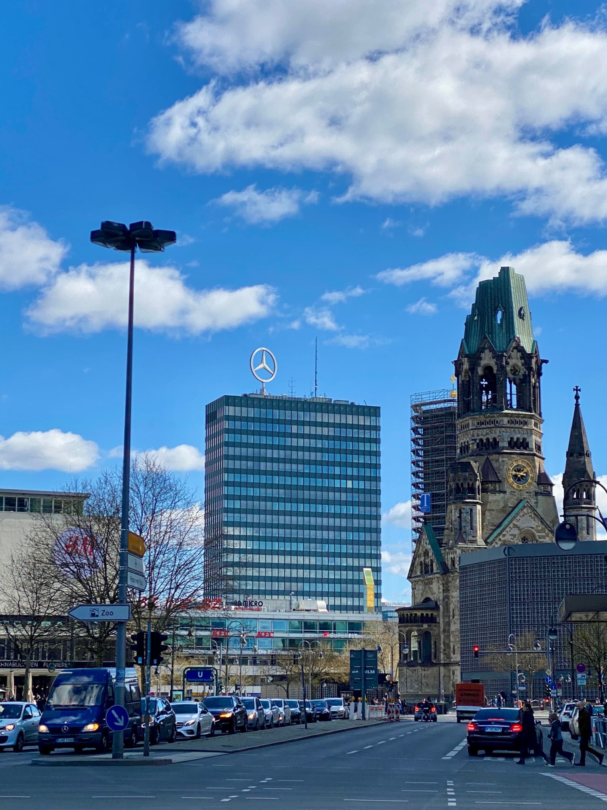 kaiser wilhelm memorial church and modern office buildings stand under a bright blue sky with traffic on a berlin city street
