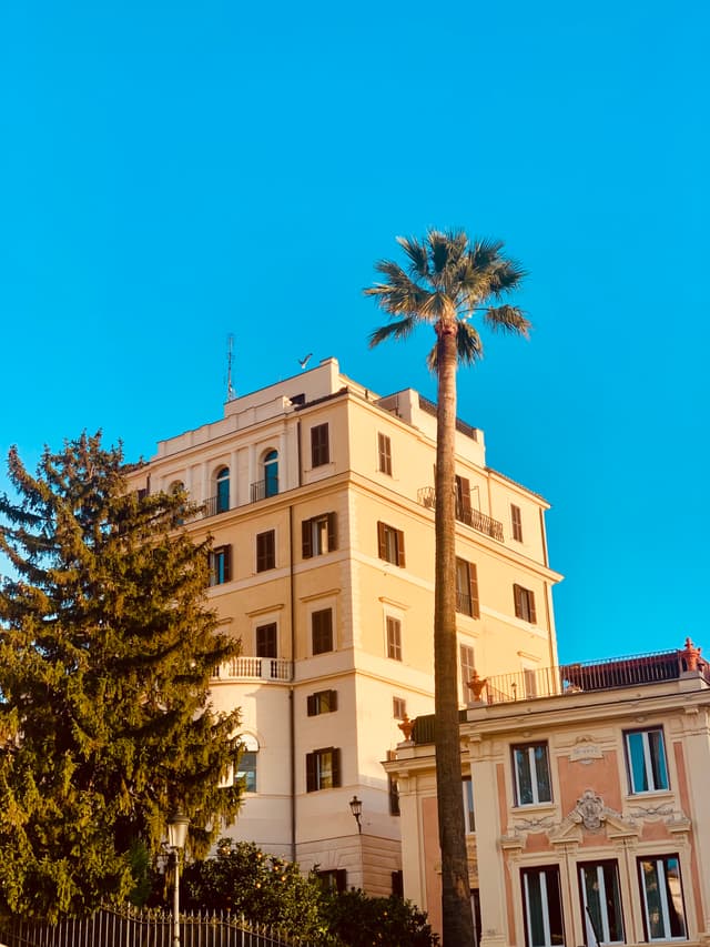 tall palm tree stands before a beige apartment building under a clear blue sky during late afternoon golden hour in rome