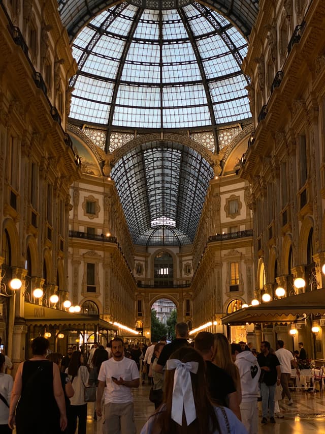 crowds walk through the galleria vittorio emanuele ii in milan under a glass dome with warm artificial lights during the evening