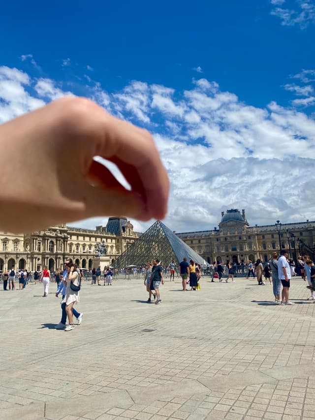 a hand pinches the louvre pyramid in paris under a bright blue sky with scattered clouds on a busy summer day
