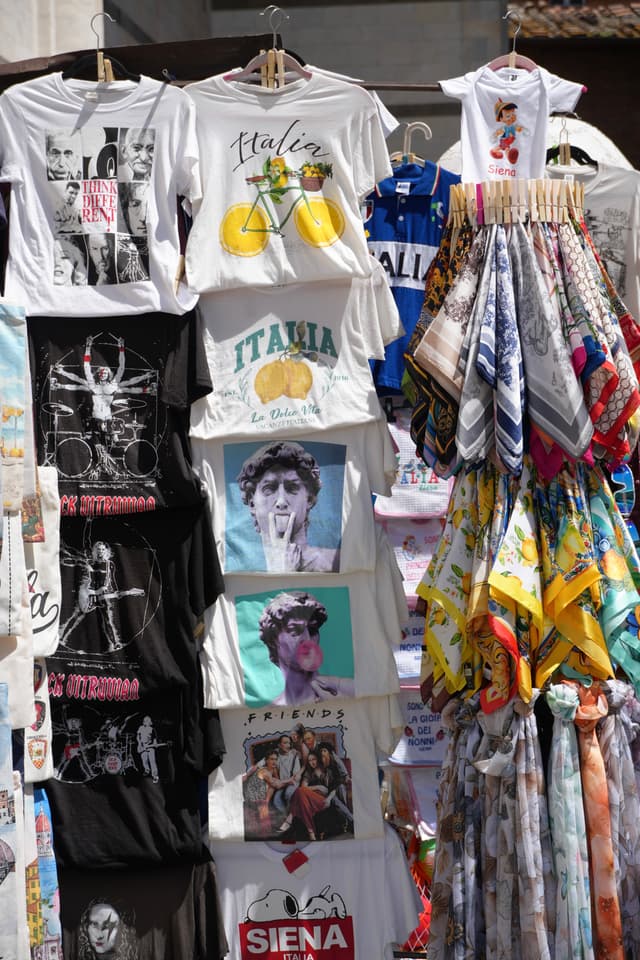 souvenir t-shirts and colorful scarves hang on a street rack in siena, italy, under bright sunlight during a busy summer day